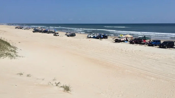 Visitors beside a line of off-road vehicles parked on South Beach at Cape Hatteras National Seashore in an archival National Park Service photograph.